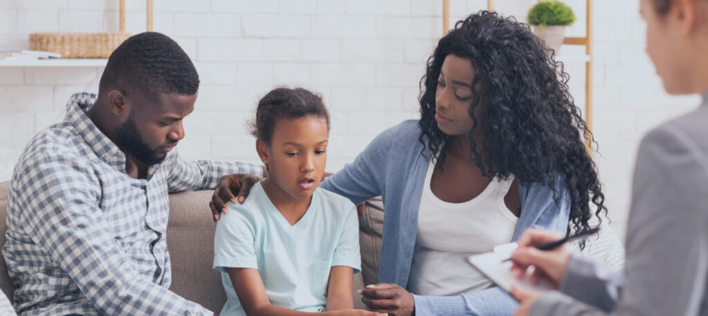 A counseling session with a mother, father, and a young girl. The three are sitting on a couch together discussing a topic.