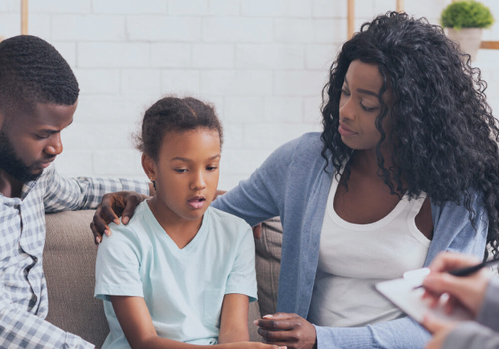 A counseling session with a mother, father, and a young girl. The three are sitting on a couch together discussing a topic.